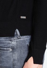 Zbliżenie na osobę w czarnym, strukturalnym swetrze z otworem na kciuk i jasnoszarych dżinsach z widoczną pętlą na pasek i nitką kieszeni.