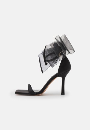 Black high-heeled sandal with a square toe, slender ankle strap, and large tulle bow accent on the heel. Smooth satin material.