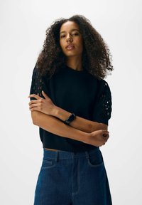 Une femme aux cheveux bouclés porte un haut noir à manches courtes avec des manches découpées, un jean bleu, et un bracelet de fitness noir à son poignet gauche.
