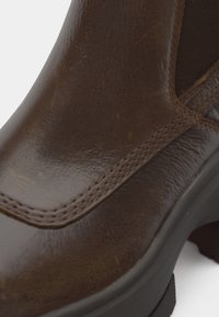 Botte de cheville en cuir marron avec une texture lisse, une forme arrondie et des détails de couture visibles, dotée d'une semelle noire contrastante pour une meilleure adhérence.