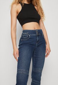 Top corto negro de canalé combinado con unos vaqueros ajustados de tiro alto en color azul que cuentan con una parte delantera abotonada y detalles de costura decorativa en las piernas.
