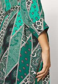 Grön mönstrad klänning med paisley- och geometriska mönster, med en texturerad tyg och knäppta manschettdetaljer.