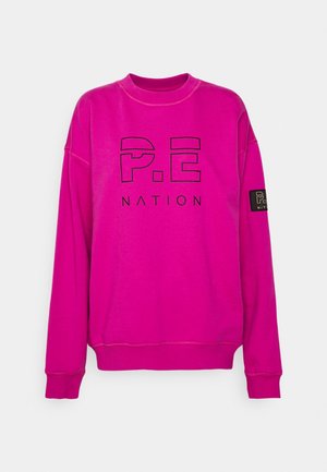 Kirkaan pinkki pitkähihainen collegepaita, jossa musta "P.E NATION" -logo edessä ja pieni musta logopatches vasemmassa hihassa.