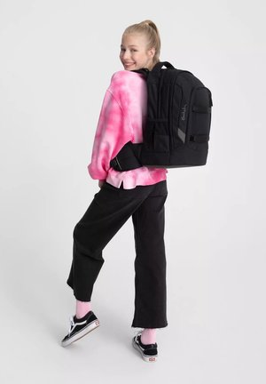 Zaino nero con più scomparti, spallacci spessi e superficie texture. Indossato sopra una felpa rosa tie-dye e abbinato a pantaloni neri.