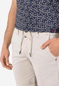 Beige pants with adjustable drawstring waist, featuring a classic cut and two side pockets. Worn with a navy blue patterned shirt.