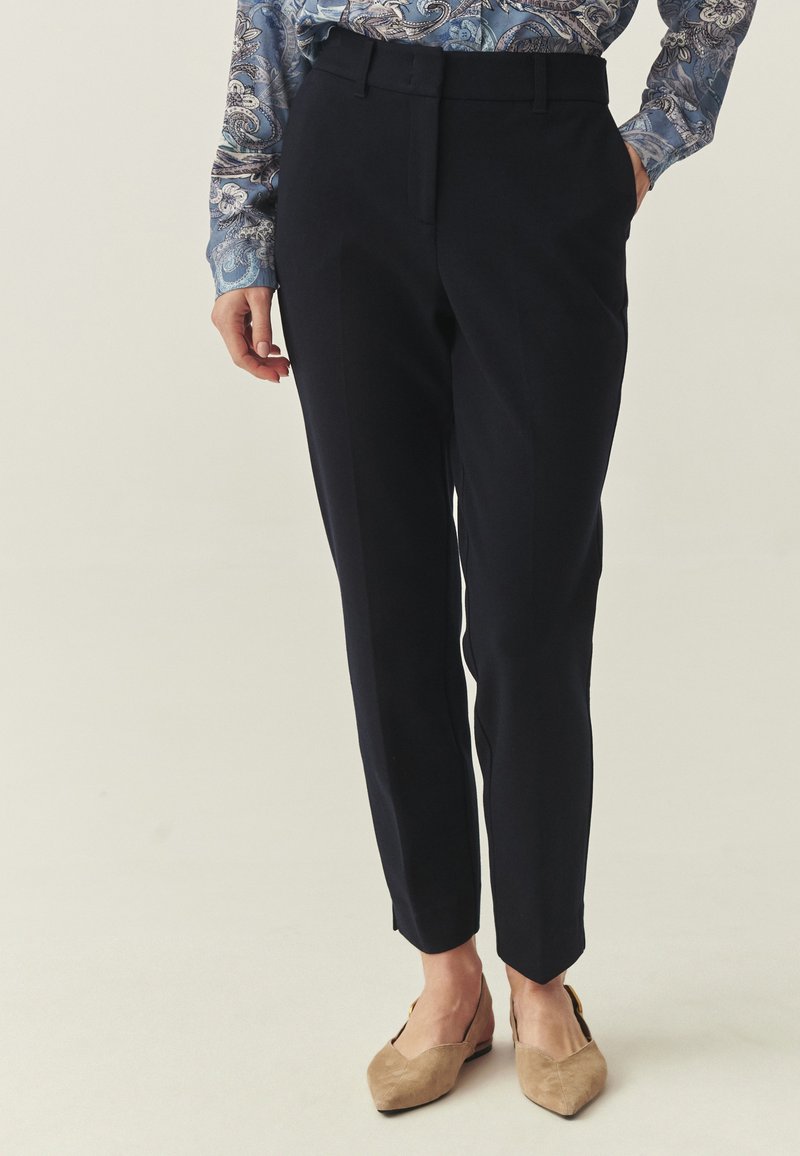 Pantalon marine ajusté, coupe slim, texture lisse, avec de discrètes pinces sur le devant, associé à des chaussures beige à bout pointu.