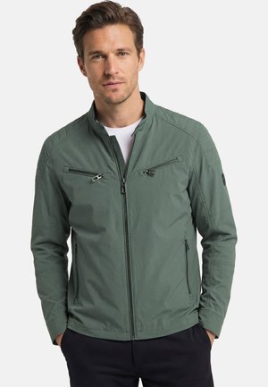 Man with short brown hair wearing a green zip-up jacket with zippered chest and side pockets, standing with hands in pockets.