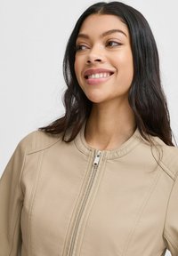 Chaqueta de piel sintética beige con escote redondo, cremallera frontal y detalles acolchados en los hombros. Textura suave y ajuste entallado.