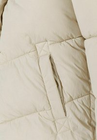 Section d'un manteau rembourré beige avec une couture diagonale et une poche avec des coutures contrastantes. Le tissu paraît lisse avec une texture douce.