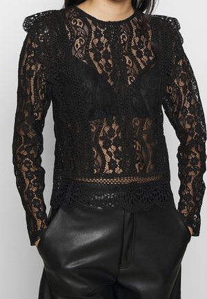 Sort gennemsigtig blonder langærmet bluse med blomsterprint, flæsedetaljer på skuldrene, båret med højtaljede sorte læderbukser med hænderne i lommerne.