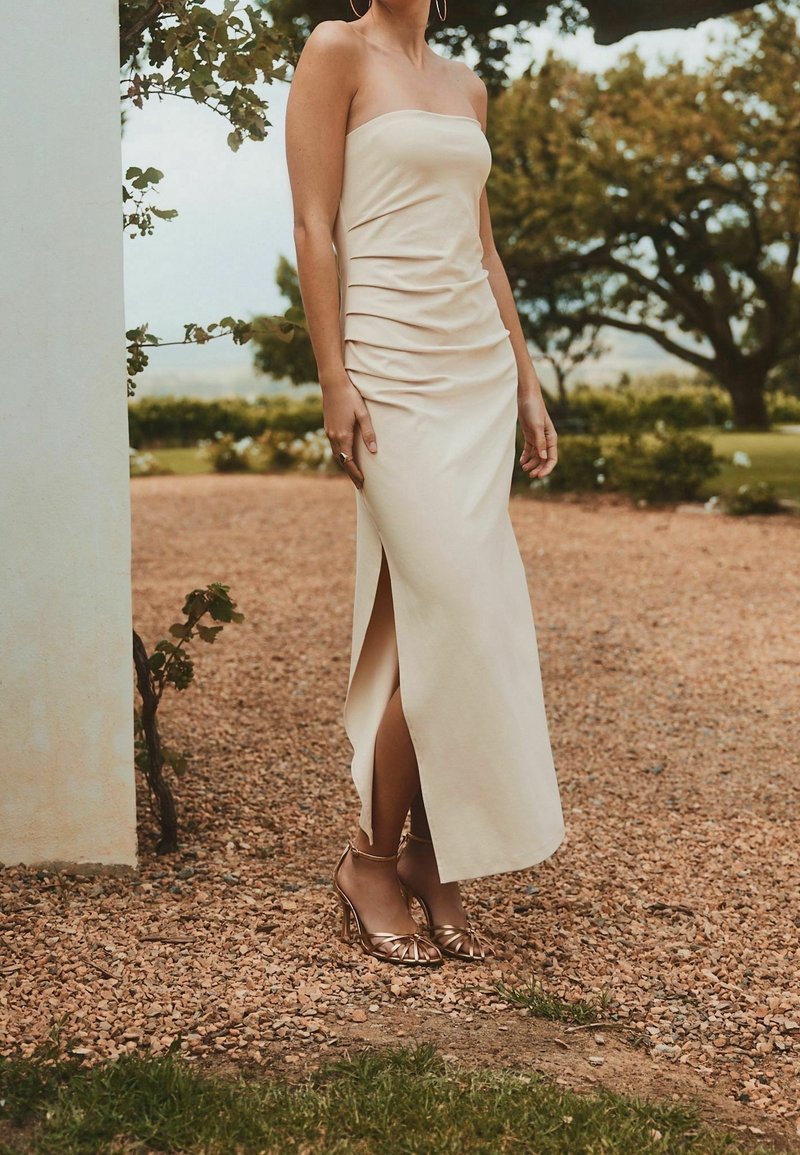 Femme portant une robe beige bustier avec une fente latérale et des sandales à talons hauts dorées, debout sur du gravier près de la verdure.