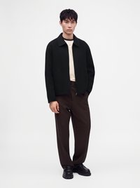 Jeune homme debout portant une veste noire, une chemise crème, un pantalon marron foncé et des chaussures noires sur un fond blanc uni.
