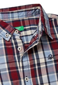 Camisa de cuadros hecha de algodón. Los colores incluyen rojo, azul marino y azul claro. Presenta un cuello abotonado y un logo bordado en el pecho.