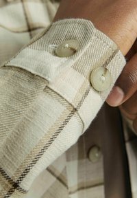 Beige geruite shirtmanchet met twee ronde, lichtgekleurde knopen. De stof heeft een zachte textuur met een geweven patroon en beschikt over bruine accentlijnen.