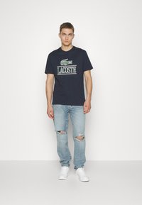 Camiseta de algodón azul marino con logo blanco de "LACOSTE" y gráfico de cocodrilo verde. Combinada con jeans claros rasgados y zapatillas blancas.