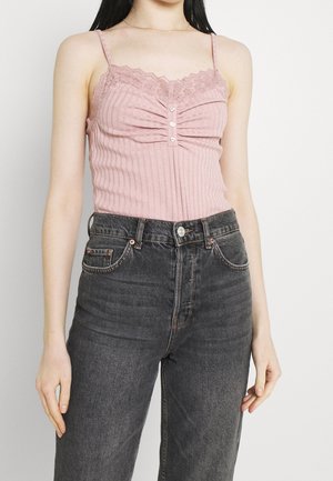 Kobieta w różowym prążkowanym topie na ramiączkach z koronkowym wykończeniem i guzikami, w połączeniu z wysokim stanem spodni jeansowych w wyblakłym czarnym kolorze, stojąca na tle białego tła.