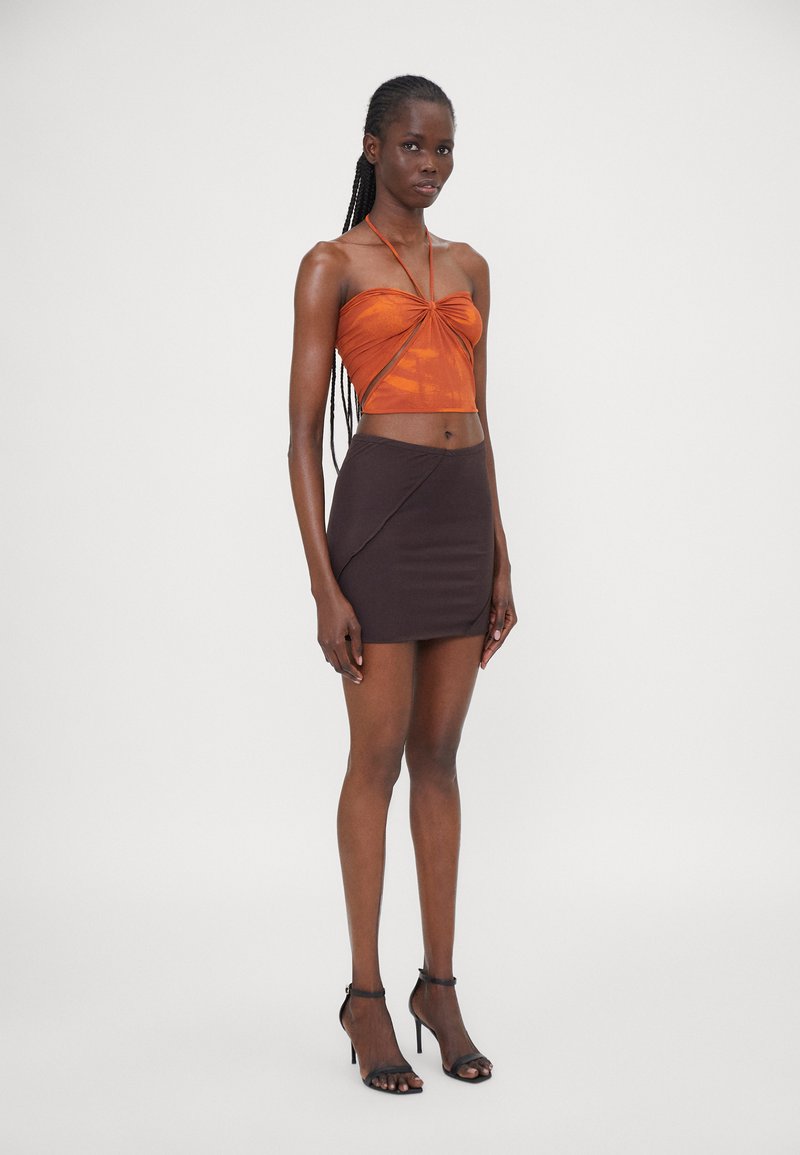 Top court en bronze avec un devant torsadé ; mini jupe noire ajustée avec un ourlet en biais ; modèle portant des talons noirs à brides ; fond blanc uni.