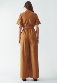 Hnědý overal se širokým páskem z hladké tkaniny, s krátkými rukávy a širokými nohavicemi, zobrazený zezadu na světlém pozadí.