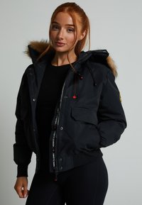 Fekete puffa dzseki prémes kapucnival és logós díszítéssel. Cipzáras és gombos záródás, oldalsó zsebek és bordázott mandzsetták jellemzik.