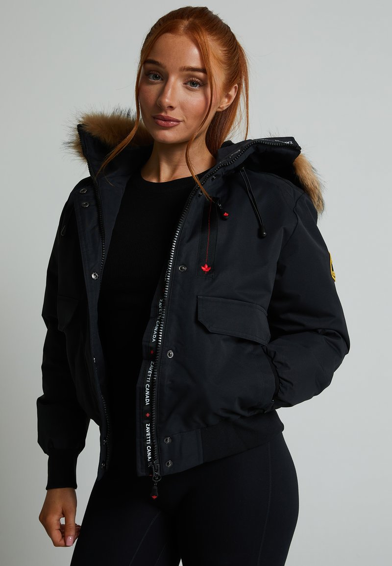 Fekete puffa dzseki prémes kapucnival és logós díszítéssel. Cipzáras és gombos záródás, oldalsó zsebek és bordázott mandzsetták jellemzik.