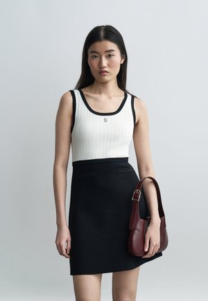 Γυναίκα με μακριά σκούρα μαλλιά, που φοράει λευκό αμάνικο τοπ και μαύρη φούστα, κρατώντας μικρό σκούρο κόκκινο τσαντάκι, μπροστά σε απλό γκρι φόντο.