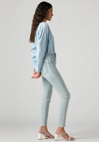 Jeans skinny de denim en un tono azul claro combinados con una camisa de manga larga en el mismo tono y sandalias de tacón cuadrado blancas. Textura suave y diseño ajustado.