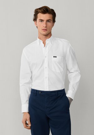 Joven con cabello castaño, vestido con una camisa blanca de manga larga abotonada y pantalones azul marino, de pie frente a un fondo liso de color gris claro.
