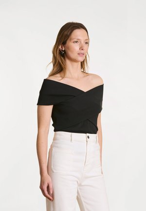 Femme aux cheveux châtain clair portant un top noir épaules dénudées et un pantalon blanc taille haute, debout devant un fond blanc uni.