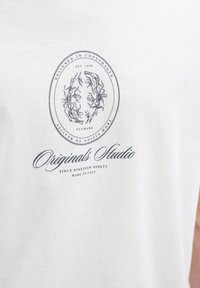Balta medvilnės marškinėliai su apvaliu grafiniu piešiniu, kuriame pavaizduota gėlių tema, ir užrašai "Suprojektuota Kopenhagoje" bei "Originalių studija" tamsiai mėlyna spalva.