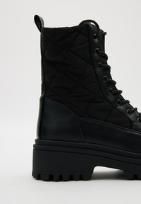 Botín negro con cordones, con una parte superior de tejido acolchado, detalles de cuero suave, suela de goma gruesa y un tacón bajo.