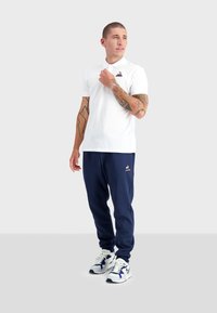 Polo blanc avec col et logo sur la poitrine, associé à un jogging marine avec un logo. Des baskets blanches et bleues complètent la tenue.