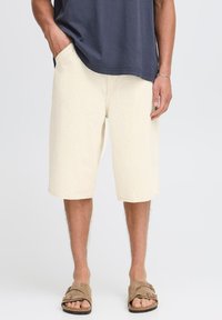 Pantalones cortos beige hasta la rodilla en una tela texturizada con bolsillos frontales, combinados con sandalias marrones y una camisa azul marino. Diseño sencillo, ajuste relajado.