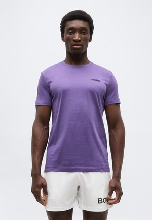 Muž v purpurovém tričku s krátkym rukávom a bielych šortkách stojaci pred jednoduchým svetlým pozadím, čelom vpřed.