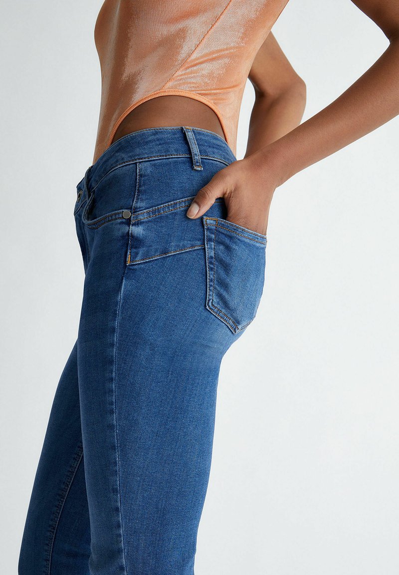 Vue de côté d'une personne portant un jean en denim bleu avec une main dans la poche arrière et un haut sans manches orange.