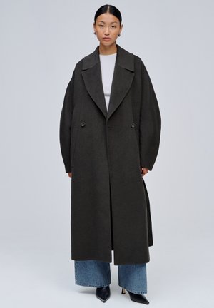 Femme aux cheveux foncés tirés en arrière porte un long manteau oversize gris foncé sur un haut blanc, un jean large bleu et des chaussures noires pointues, debout droite.