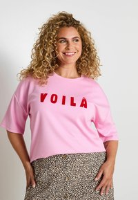Camiseta de manga corta rosa con "VOILÀ" en letras rojas, combinada con una falda beige de estampado de leopardo con detalles de botones. Material suave, corte holgado.