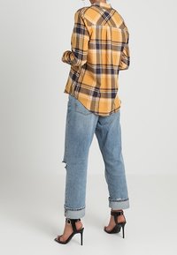 Persona che indossa una camicia a quadri gialla, jeans azzurri con risvolto e un piccolo strappo, e sandali neri con tacco a stiletto e cinturino alla caviglia, in piedi davanti a uno sfondo neutro.