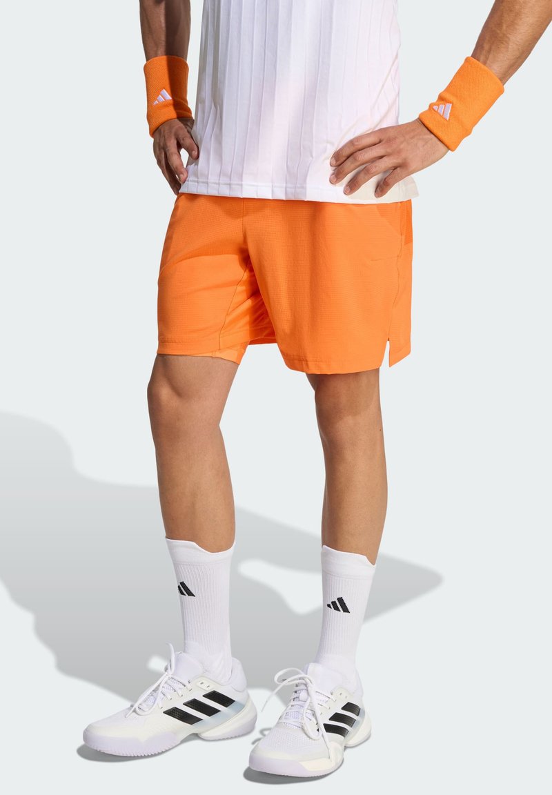 Λευκό αθλητικό μπλουζάκι με ραβδώσεις, πορτοκαλί σορτς με υφή, λευκές κάλτσες με μαύρες ρίγες και λευκά παπούτσια με μαύρες λεπτομέρειες. Πορτοκαλί βραχιόλια στον καρπό.