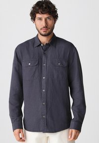 Dunkelgraues Hemd mit langen Ärmeln, zwei Brusttaschen und einer weichen Textur. Doppelte Knöpfe an den Manschetten und ein klassischer Kragen.