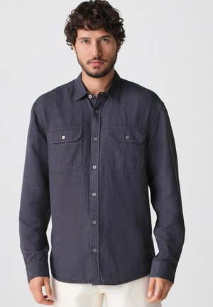 Camicia button-up grigia scura con maniche lunghe, due tasche frontali e una texture morbida. Doppi bottoni ai polsini e colletto classico.