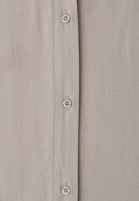 Tissu de chemise beige avec trois boutons blancs alignés verticalement sur la patte de boutonnage, montrant des coutures détaillées et une texture lisse.