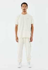 Camisa de manga corta color crema y joggers a juego, hechos de una tela suave con costuras grises en contraste y detalles del logo, combinados con zapatos blancos.