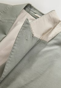 Col de veste gris clair texturé avec doublure intérieure beige, montrant les détails de la couture et la boutonnière sur le revers.