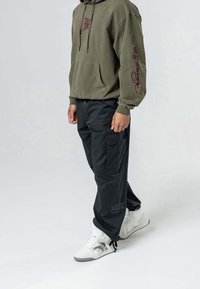 Persoon in een olijfgroene hoodie met rode tekst op de mouw, zwarte cargobroek met logo bij de enkel, en witte hoge sneakers met zwarte accenten.