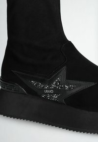 Stivaletto nero in suede con suola a zeppa. Presenta un accento a forma di stella nera testurizzata ricoperta di glitter e un logo ben visibile.