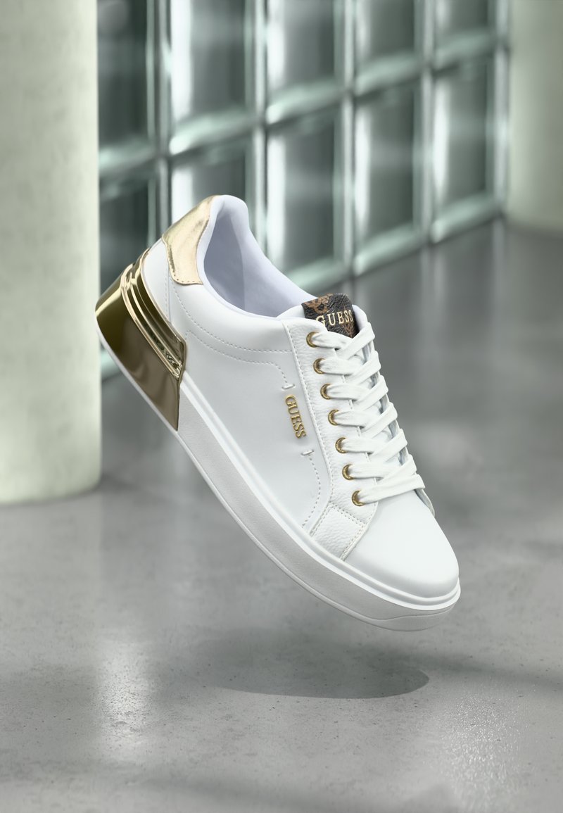 Sneaker bianco con dettagli dorati e branding "GUESS" esposto su uno sfondo di parete in vetro riflettente e pavimento in cemento.