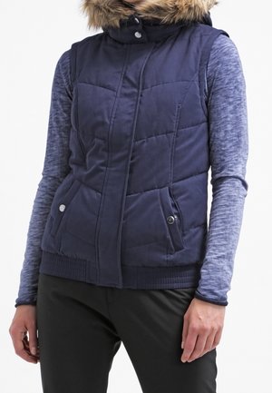 Personne portant une veste matelassée bleu marine avec capuche bordée de fourrure par-dessus une chemise bleu chiné à manches longues et un pantalon noir.