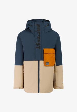 Garçon portant une veste d'hiver color block bleu marine et beige, un pantalon, un bonnet orange et des bottes noires, debout les mains dans les poches.