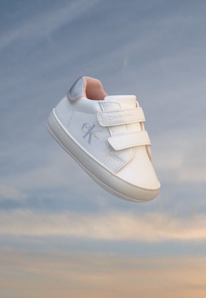 Baskets blanches Calvin Klein avec deux brides Velcro flottant sur un fond de ciel nuageux, détail du talon rose clair visible.