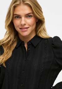 Mustvalge kootud pluus, millel on nööpidega ees, krae ja tekstuuriga muster, kus on väiksed lõiked ja puhvärv.